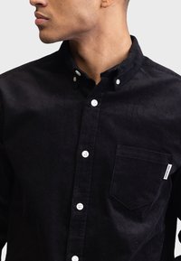 Camicia nera in corduroy con bottoni bianchi, piccola tasca sul petto e una finitura testurizzata. Il colletto è di tipo button-down.