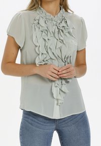 Blouse vert clair à manches courtes avec un devant froncé et un col froncé. Tissu lisse, fermeture à boutons, silhouette ajustée, associé à un jean bleu.