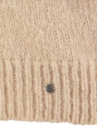 Beige gestrickter Stoff mit einer gerippten Struktur, versehen mit einem kleinen silbernen Metalletikett mit eingravierter Schrift. Weich und warm im Aussehen.