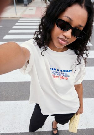 Young woman wearing black sunglasses and a white printed T-shirt takes a selfie on a street crosswalk holding a yellow bag.