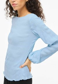 Top a maniche lunghe di colore azzurro chiaro realizzato in tessuto strutturato, con scollo rotondo, orlo arricciato e maniche a sbuffo.