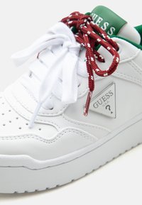 Weißer Guess-Sneaker mit grünem Kragen, rot-weiß gemusterten Schnürsenkeln und weißen Schnürsenkeln, die zu Schleifen gebunden sind, mit metallischem Guess-Logo an der Seite.