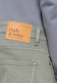 Πράσινο τζιν παντελόνι με καφέ δερμάτινη ετικέτα με ανάγλυφο το λογότυπο "Only & Sons" και πορτοκαλί ραφές. Γκρι ύφασμα ορατό πάνω από τη μέση.