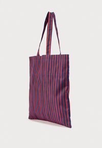 Τσάντα ώμου από ύφασμα με κάθετες ρίγες σε κόκκινο, μπλε και μαύρο χρώμα, με δύο μακριές λαβές, που κρέμεται πάνω σε απλό λευκό φόντο.