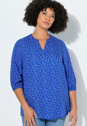 Kvinde med krøllet hår iført en blå bluse med små hvide hjerte-mønstre og trekvartærmer, stående mod en hvid baggrund.