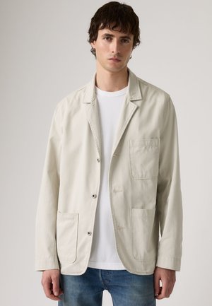 Homme portant une veste décontractée beige clair avec trois poches avant, sur une chemise blanche et un jean bleu, debout devant un fond uni.