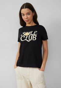 Zwarte katoenen t-shirt met grote witte tekst "SALE CLUB" en een zon grafiek. Korte mouwen en een relaxed fit, gecombineerd met lichte broek.