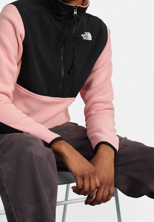 Persoon die een roze en zwarte North Face fleece trui draagt met een rits op de borst, zittend met handen rustend op een donkere broek.