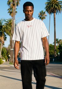 Muž kráčajúci vonku, oblečený v bielom tričku s červeným podpisovým logom a čiernych nohaviciach, v pozadí palmy a jasná obloha.