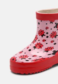 Rosa Gummiregenstiefel mit roten Akzenten, versehen mit einem Muster aus Marienkäfern und schwarzen Punkten. Glatte Textur und geriffelter Sohle für besseren Halt.