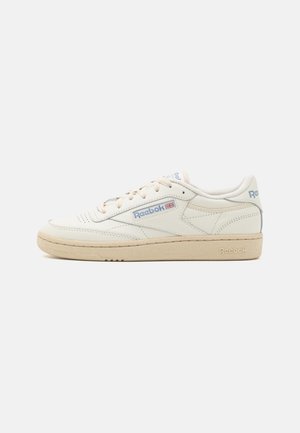 Witte leren sneakers met beige rubberen zolen, voorzien van blauwe accenten en een zichtbaar merklogo aan de zijkant. Rond neusontwerp.