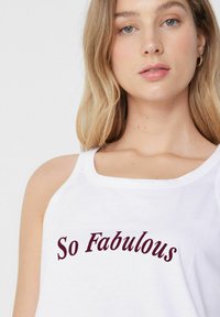Bílý bezrukávový top s nápisem "So Fabulous" v bordó kurzívě. Hladká bavlněná tkanina a kulatý výstřih.
