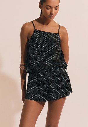 Frau trägt schwarzen ärmellosen gepunkteten Oberteil und passende Shorts, steht vor einem einfarbigen beigen Hintergrund und schaut nach unten.