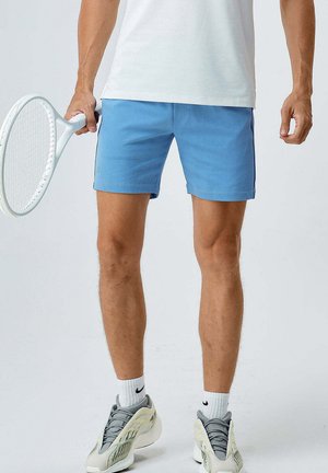 Shorts sportivi blu con una fascia elastica in vita, indossati con una maglietta bianca e scarpe da ginnastica grigie. Tiene in mano una racchetta da tennis bianca.