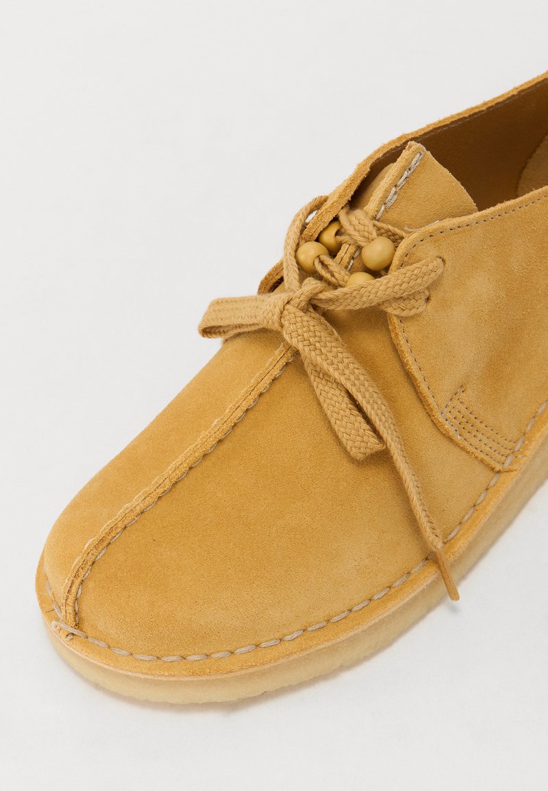 Suède casual schoen in mosterdgeel met een ronde neus, voorzien van een knoopkoord en houten kralenaccenten, gestikte details rond de zool.