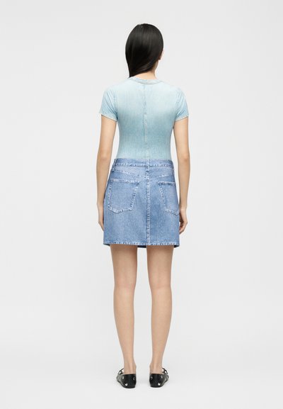 Γυναίκα με μακριά σκούρα μαλλιά φοράει κοντομάνικη γαλάζια μπλούζα και δίχρωμη τζιν φούστα με πίσω τσέπες, στέκεται με την πλάτη προς τα εμπρός.