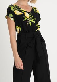 Top negro con estampado floral en amarillo y verde, escote en V, mangas cortas; combinado con pantalones de tiro alto negros con un cinturón atado.