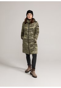 Lang olijfgroen pufferjas met een hoge kraag, ritssluiting, zijzakken en textuurpanelen. Draagt met zwarte jeans en bruine enkellaarsjes.