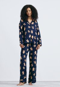 Vrouw staat blootsvoets in een marineblauw pyjama-set met beige peperkoeken mannen, rendieren en sterrenpatronen tegen een effen witte achtergrond.