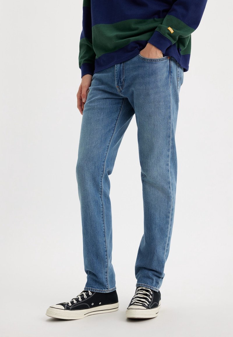 Personne portant un jean bleu, des baskets noires et blanches, et un t-shirt à manches longues bleu et vert, debout avec une main dans la poche.