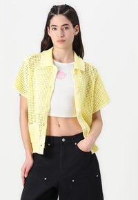 Gul stickad skjortjacka med korta ärmar, öppen väv, krage, knappar framtill och ficka; bärs över en vit croptopp och svarta byxor.