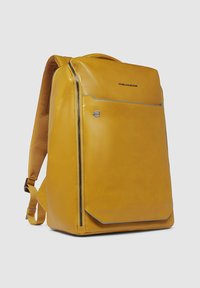 Mochila de cuero amarilla con una textura suave, forma rectangular, bolsillo delantero con cremallera y detalle de costura azul en los bordes. Correas ajustables.