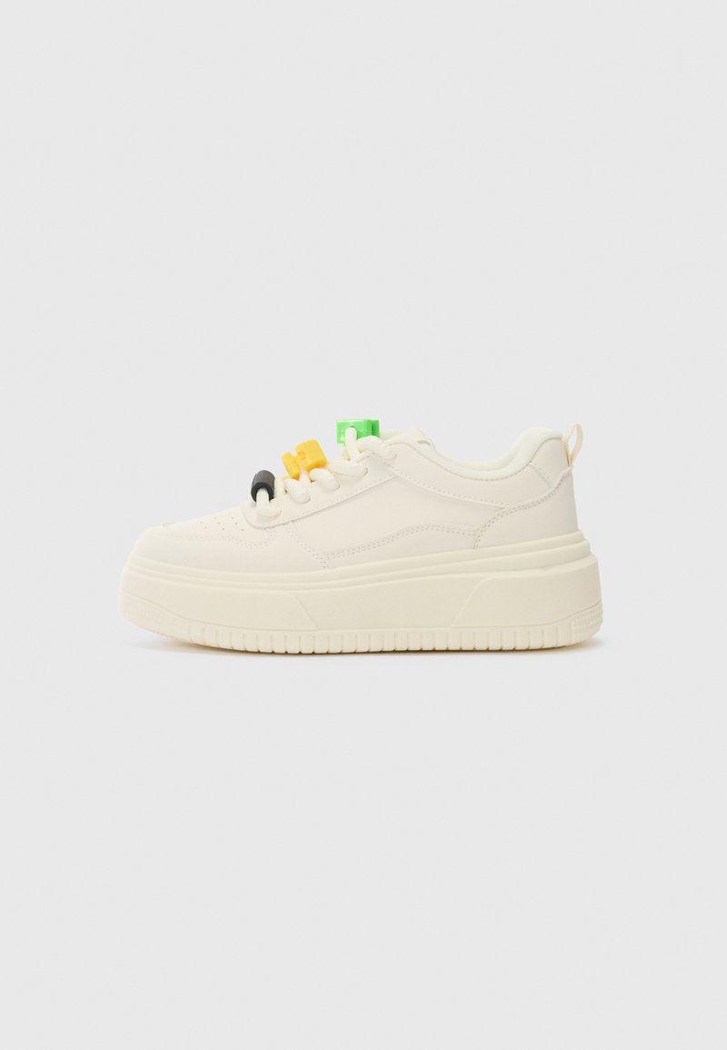 Weißer Sneaker mit dickem Sohlenprofil, strukturiertem Obermaterial und kontrastierenden Schnürsenkeln in Grün, Gelb und Schwarz. Runde Zehenform und Ziehlasche an der Rückseite.