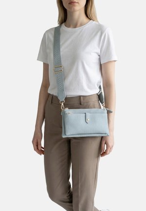 Donna con maglietta bianca e pantaloni marroni che porta una borsa a tracolla azzurro chiaro con tracolla larga decorata, su sfondo bianco.