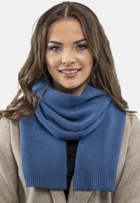 Căciulă tricotată albastră cu o textură netedă și margini cu fuste, așezată în jurul gâtului, asociată cu o haină gri deschis.