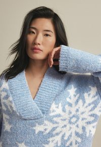 Pullover oversize di colore azzurro chiaro con scollatura a V, realizzato in tessuto a maglia testurizzato. Presenta motivi di fiocchi di neve bianchi sul davanti.