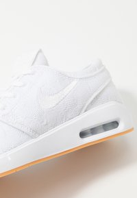 Vit atletisk sko med strukturerad tygöverdela, tonad swoosh-logotyp, vit gummisula och synlig luftkudde. Orange detalj på yttersulan.