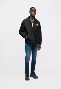 Μαύρο νάιλον μπουφάν με κόκκινο λογότυπο στο στήθος, συνδυασμένο με μπλε τζιν παντελόνι και μαύρα αθλητικά παπούτσια. Το μπουφάν έχει χαλαρή εφαρμογή και κούμπωμα με κουμπιά.