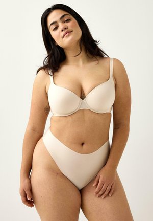 Kvinde med langt mørkt hår iført beige bh og trusser med høj talje, stående foran en ensfarvet lys baggrund, med tatoveringer på armene.