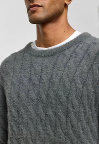 Grijze kabelgebreide trui van zacht, textuurmateriaal. Heeft een brede hals en een gelaagd uiterlijk met een wit shirt eronder.