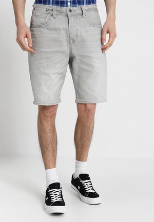 Homme portant un short en denim gris clair, des chaussettes blanches et des baskets noires à lacets blancs, debout devant un fond blanc uni.