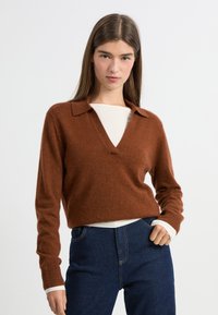 Brązowy sweter z mieszanki wełny z dekoltem w kształcie V i kołnierzykiem. Posiada długie rękawy oraz luźny krój, noszony na białej bluzce.