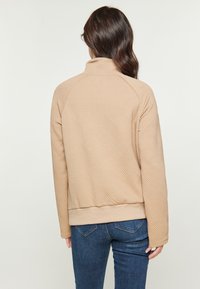 Beige steppelt pulóverdzseki magas gallérral, geometrikus mintával, bordázott textúrával és fitted derékkal, farmernadrággal párosítva.