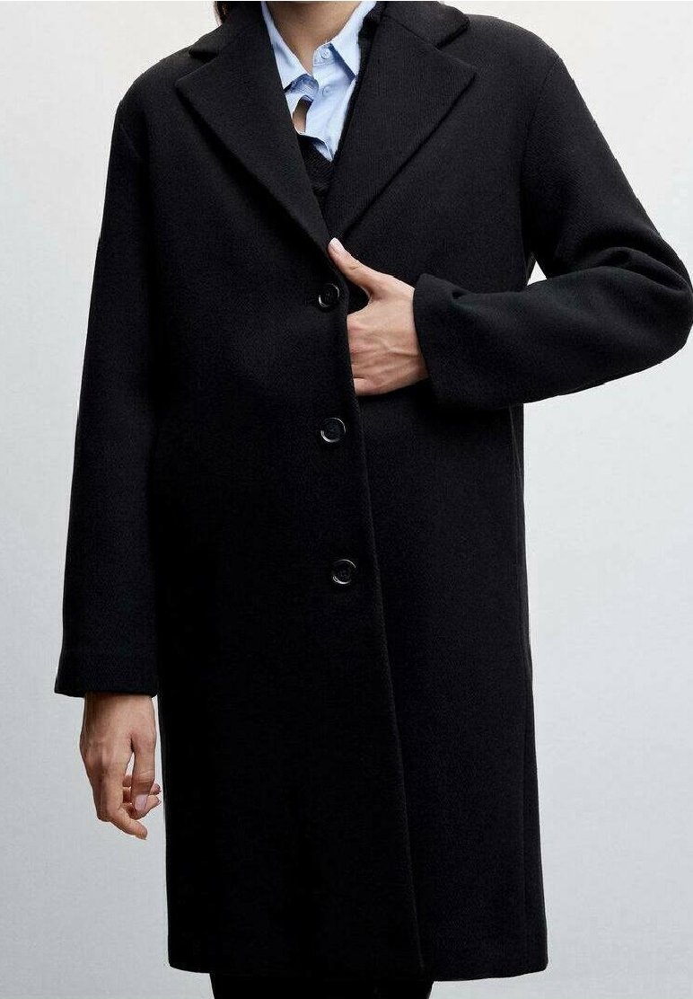 Personne portant un manteau noir boutonné par-dessus une chemise bleu clair à col, tenant le manteau fermé à la poitrine avec une main.