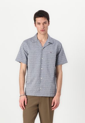 Chemise à manches courtes, boutonnée, en tissu bleu clair avec un motif géométrique, texture douce, col cranté, et petit logo sur la poitrine gauche.