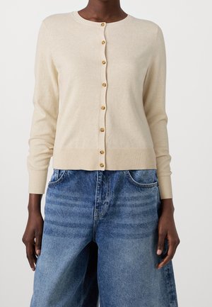 Person iført en beige knappet cardigan med ribbet kant og manchetter kombineret med løstsiddende blå jeans.