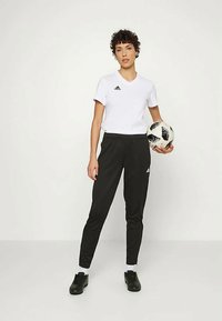 Cămașă sport albă cu mânecă scurtă, cu logo-ul Adidas negru, asociată cu pantaloni negri îngustați. Minge de fotbal neagră în mână, încălțăminte sport neagră la picioare.