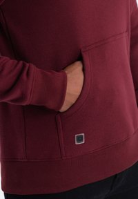 Felpa bordeaux con tasca a marsupio, con polsini e orlo ribattuti. Presenta un piccolo logo quadrato vicino alla tasca. Materiale morbido e texturizzato.