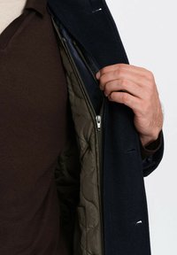 Cappotto in misto lana con esterno blu navy, caratterizzato da un rivestimento interno in verde oliva trapuntato e chiusura con zip, che mette in risalto cuciture dettagliate e due tasche esterne.
