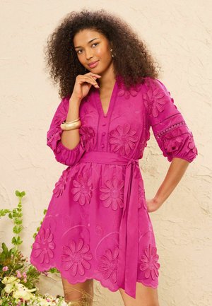 Femme aux cheveux bouclés portant une robe rose vif brodée de fleurs avec des manches bouffantes et une ceinture à la taille, posant devant un mur beige texturé.