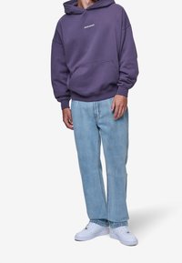 Fialová mikina s kapucí a přední kapsou, kombinovaná s světle modrými džíny a bílými teniskami. Neformální, volný střih.