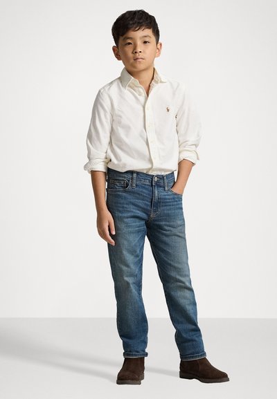 Balta krekla ar pogām, uz kuras ir neliela logo, zili denima džinsi un brūni zamša zābaki. Apģērbs ir brīvs un ar klasisku dizainu.