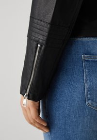 Black faux leather jacket with quilted shoulder details and silver zipper at the cuff. Worn with blue denim jeans.