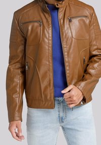 Giacca di pelle marrone con colletto, tasche con zip, costine orizzontali sulle spalle e una finitura liscia, indossata sopra un maglione blu.