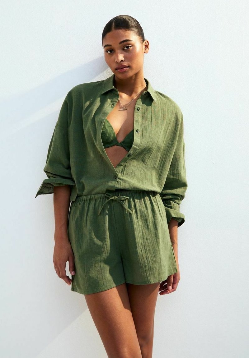 Chemise boutonnée vert olive portée par-dessus un haut de bikini assorti et un short, avec une coupe décontractée et une taille à cordon, en tissu texturé.