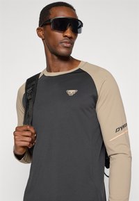 Hosszú ujjú póló fekete testtel, bézs ujjakkal, logóval a mellkasán és felirattal az ujjon. Nagy fekete napszemüveggel és hátizsákkal kiegészítve.